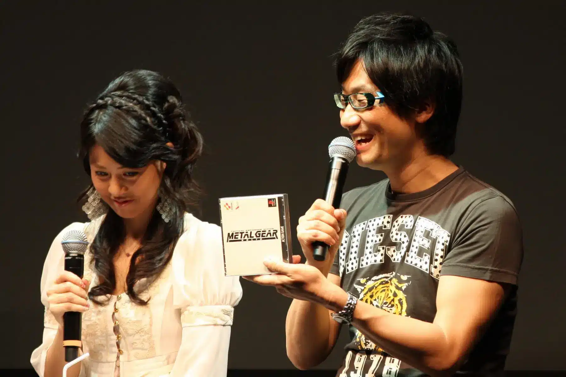 Hideo Kojima holding a Metal Gear game case while speaking into a microphone.
